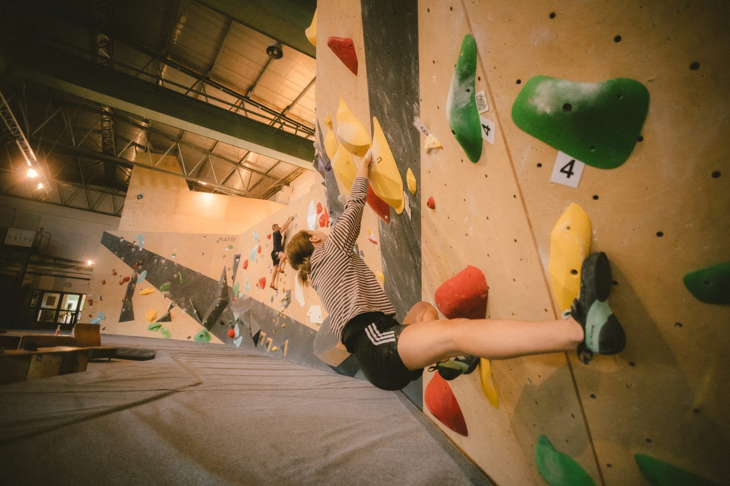 Boulderin im weiten Spreizschritt, dahinter die Halle