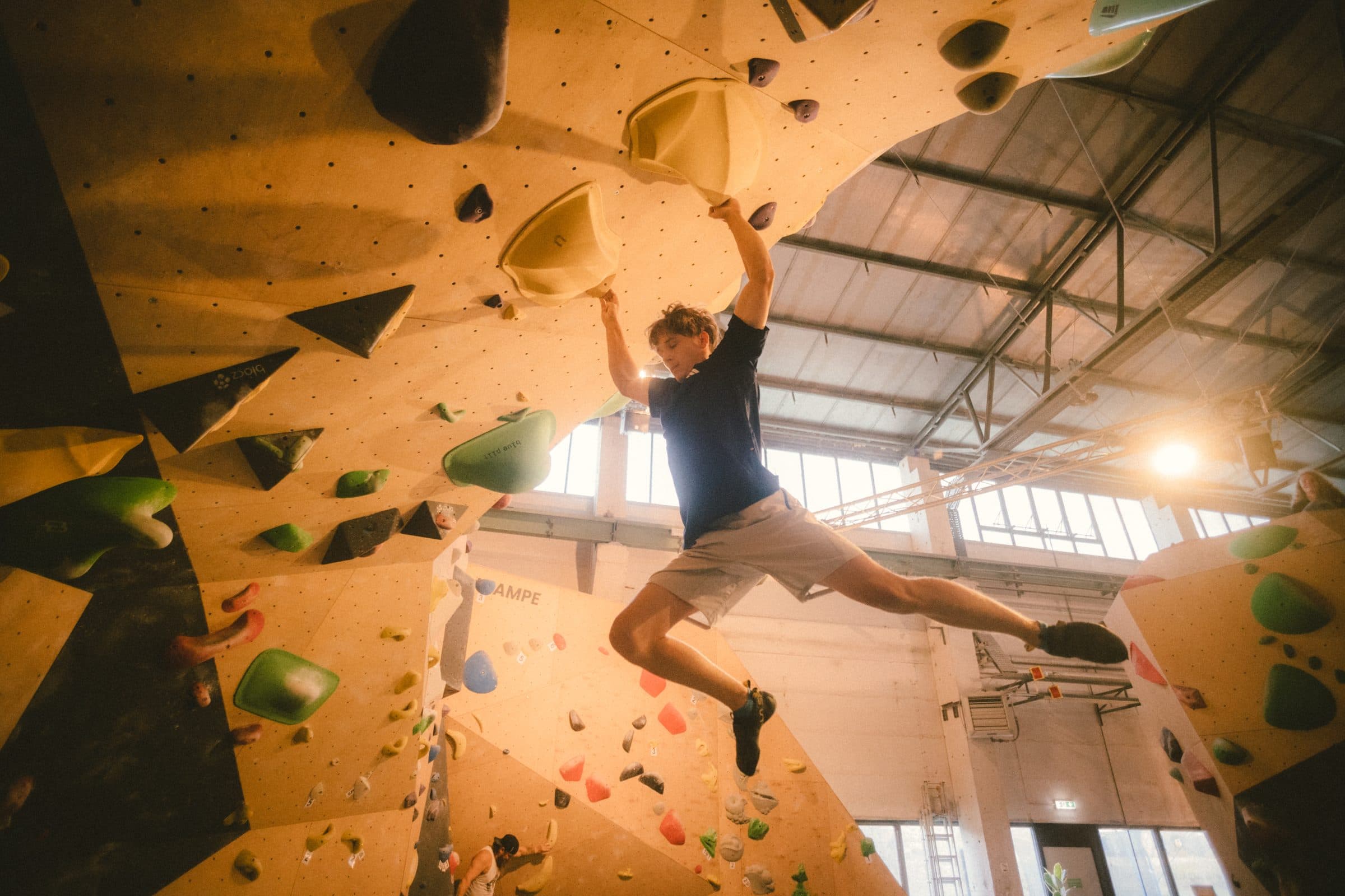 Boulderer schwingt im Überhang frei, dahinter die Fensterfront der Halle